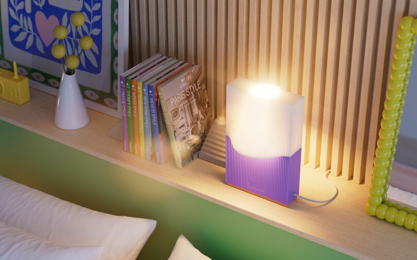 A bedside table with a colorful lamp, books, and decorative items, including a framed picture and a vase with yellow flowers. The lamp is turned on, casting a warm light