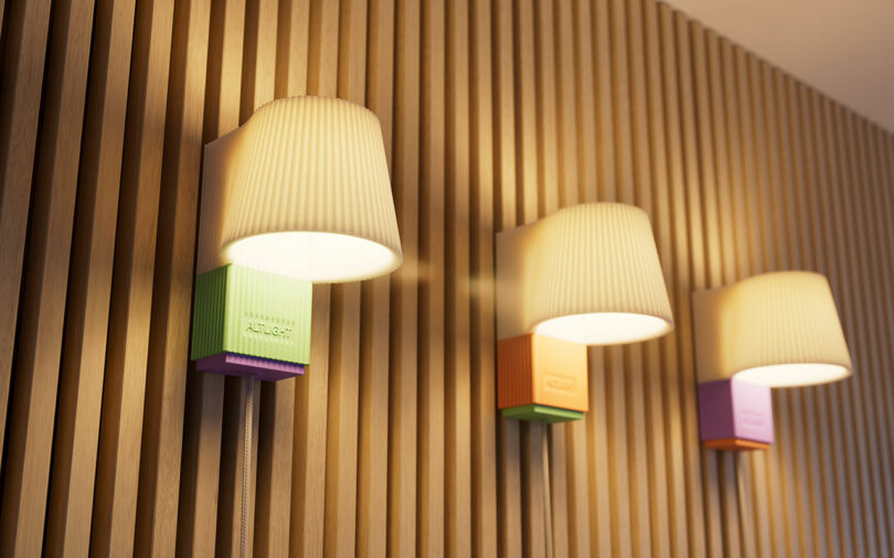 Three wall-mounted lights with pleated white lampshades are fixed to a wood paneled wall. Each light has a colored base in green, orange, and purple respectively