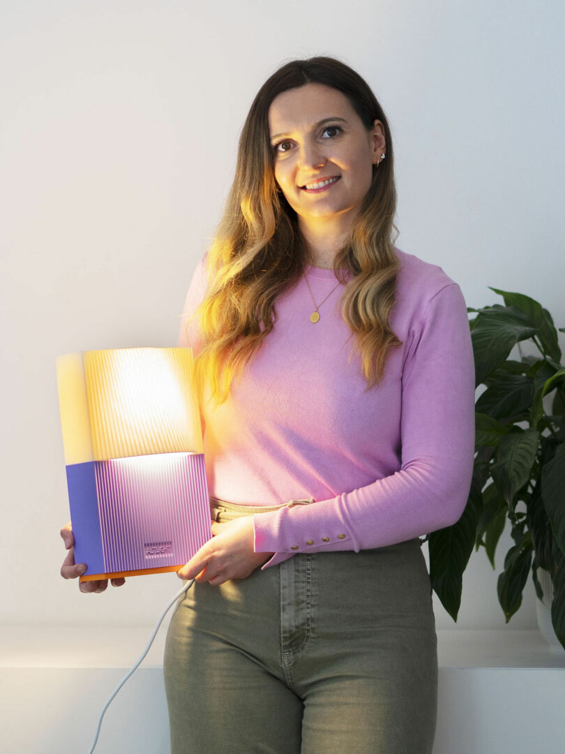 A woman in a pink sweater and green pants holds a rectangular lamp with purple and yellow sections while standing indoors near a plant