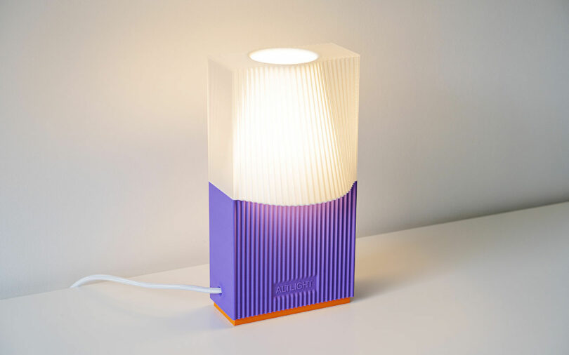 Modern tabletop lamp with a white, pleated shade and a purple and orange rectangular base, placed on a white surface