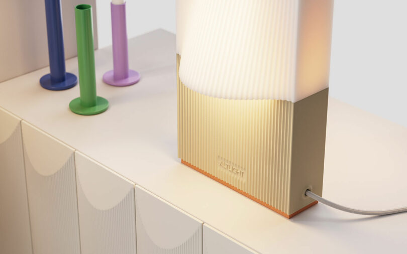 A modern lamp with a ribbed, two-toned design emits a warm light. It is placed on a white cabinet next to colorful candle holders in green, blue, and purple