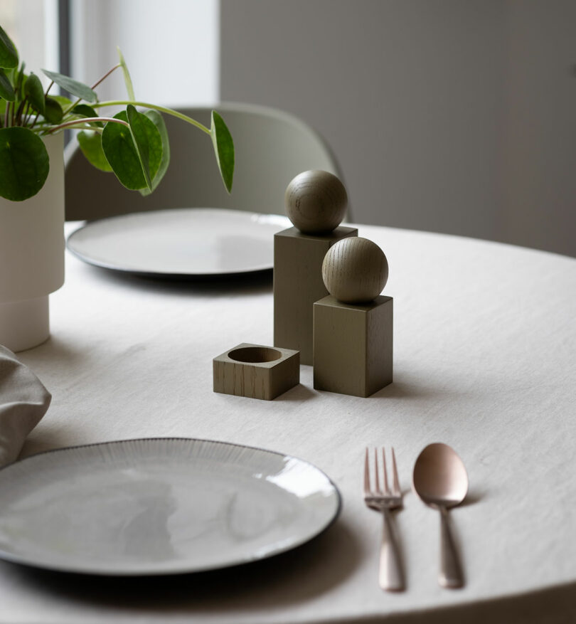 A dining table with two place settings, each with a plate, fork, knife, and spoon. There are two wooden decorative sculptures and a potted plant near the edge of the table.