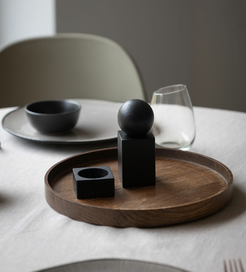 A wooden tray holds a black sphere on a black cube and a black cylindrical holder, on a table set with dishes, a bowl, and a glass