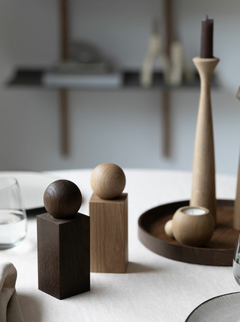 Two wooden sculptures with spheres on top sit on a round table. In the background, a wooden tray with a tall candleholder and small round objects are visible on a blurred shelf.