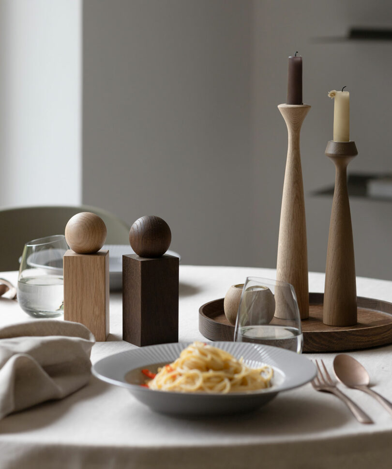 A table setting with a plate of spaghetti, two glasses of water, wooden candle holders with candles, and decorative wooden objects in the background