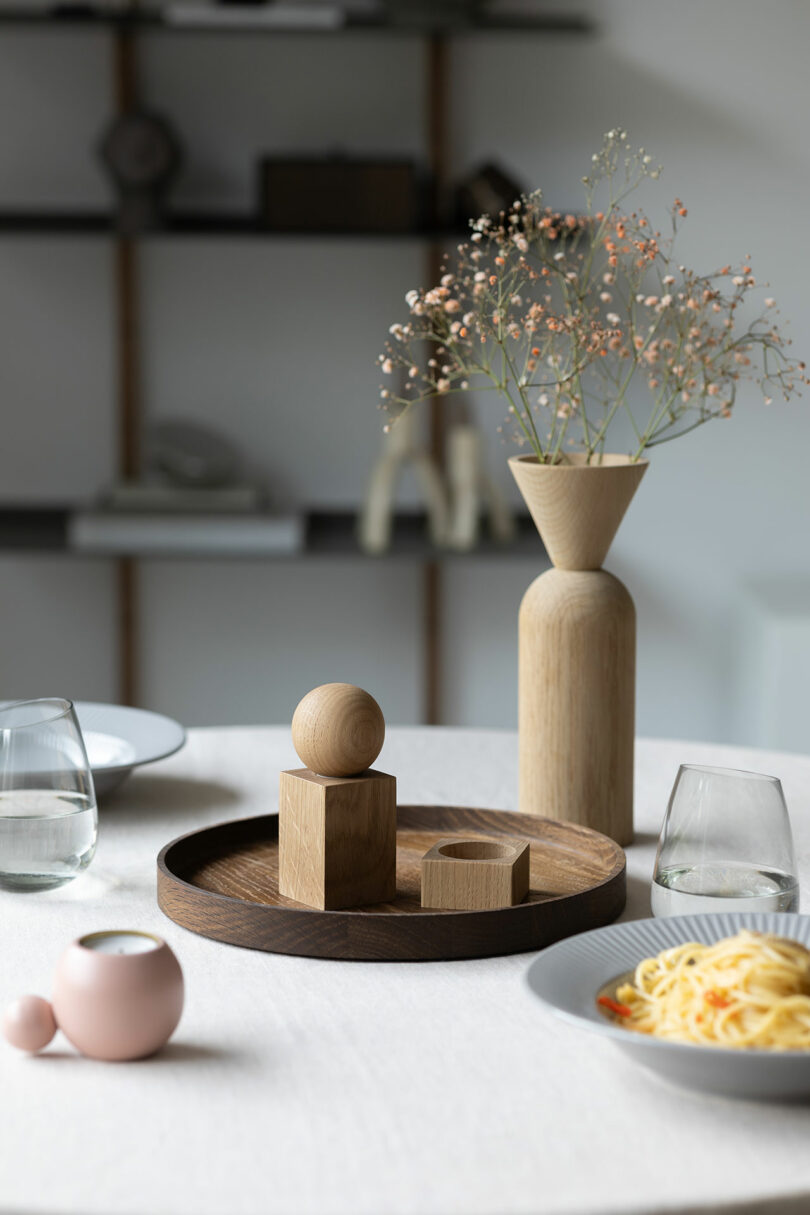 A round table with plates of pasta, two glasses of water, wooden salt and pepper shakers, and a vase with dried flowers