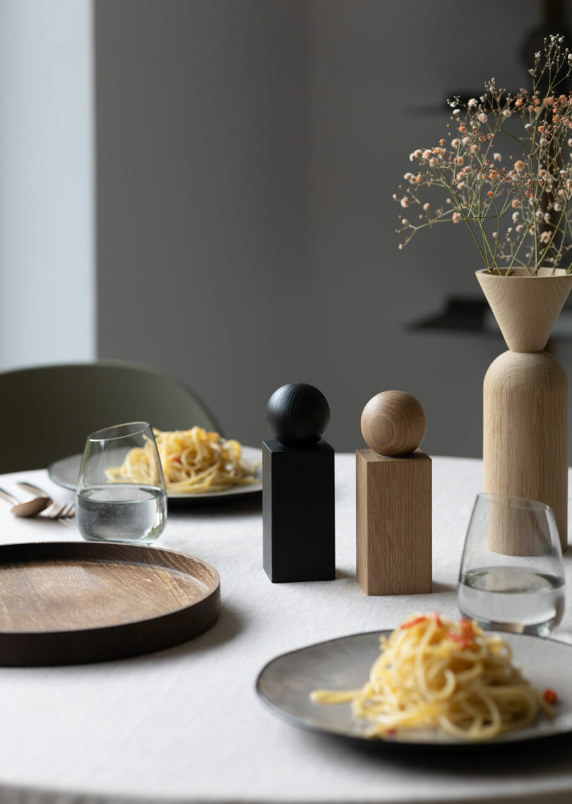 A round table with plates of pasta, two glasses of water, wooden salt and pepper shakers, and a vase with dried flowers