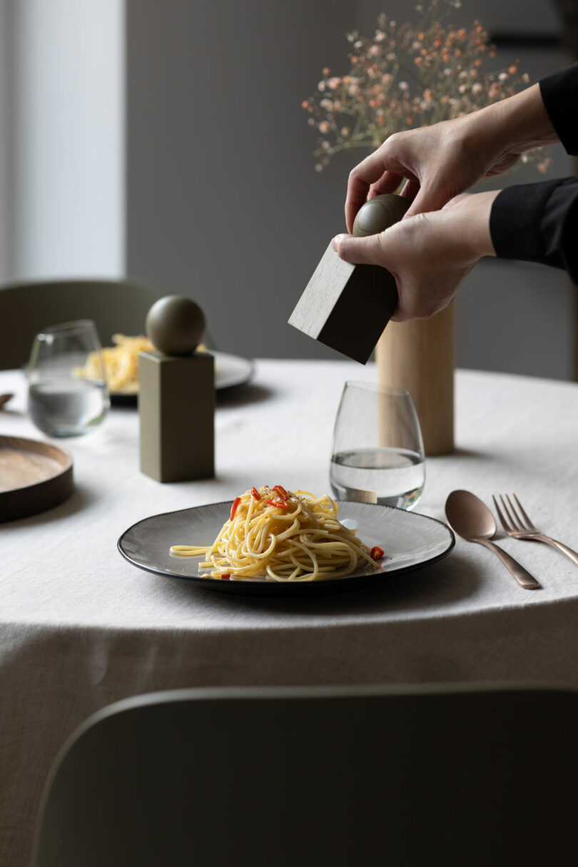 Person grinding pepper onto a plate of spaghetti on a table set for a meal, with glasses of water, cutlery, and centerpieces in the background