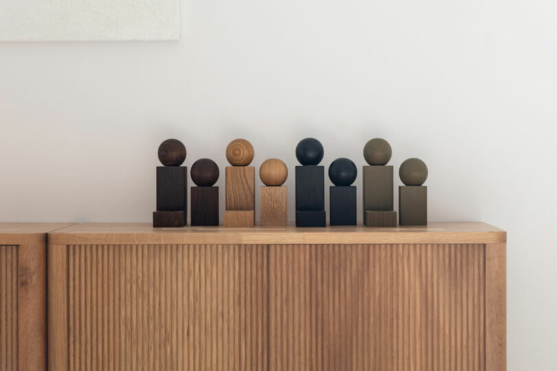 A series of wooden geometric sculptures with round and rectangular shapes in various wood tones are arranged on top of a wooden cabinet against a plain white wall