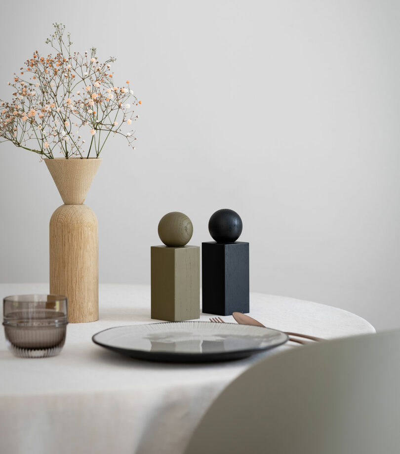 A minimalistic table setting with a round, white tablecloth, a clear drinking glass, a white plate with cutlery, a wooden vase with flowers, and two decorative blocks with spherical tops