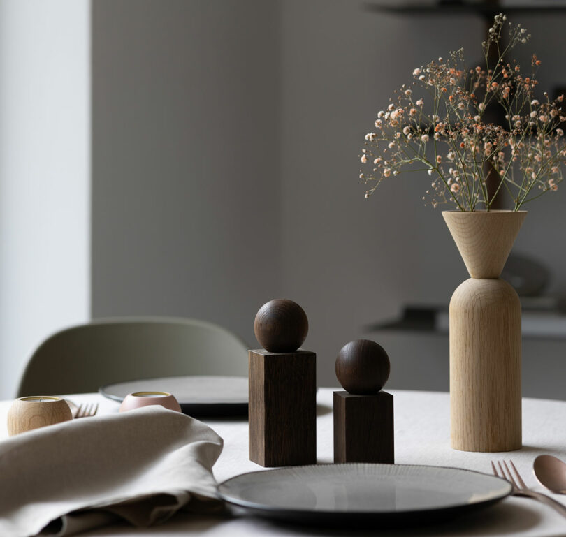 A dining table set with two black plates, folded beige napkins, a wooden vase with flowers, two wooden sculptures, and utensils on a light-colored tablecloth. A green chair is in the background