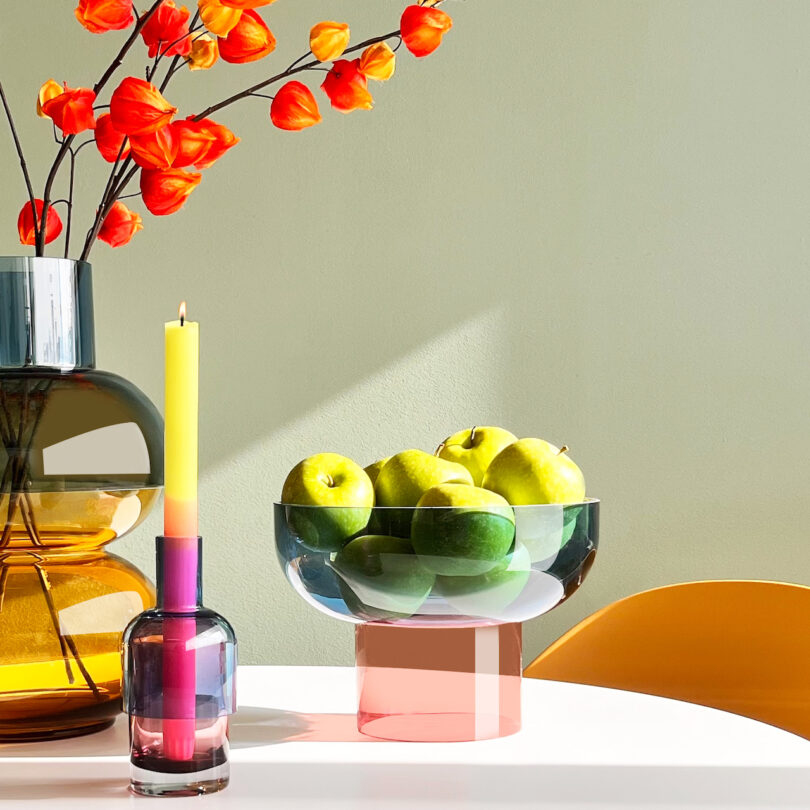 A table holds a vase with orange flowers, a two-tone candle in a holder, and a glass bowl filled with green apples. A yellow chair is partially visible on the right