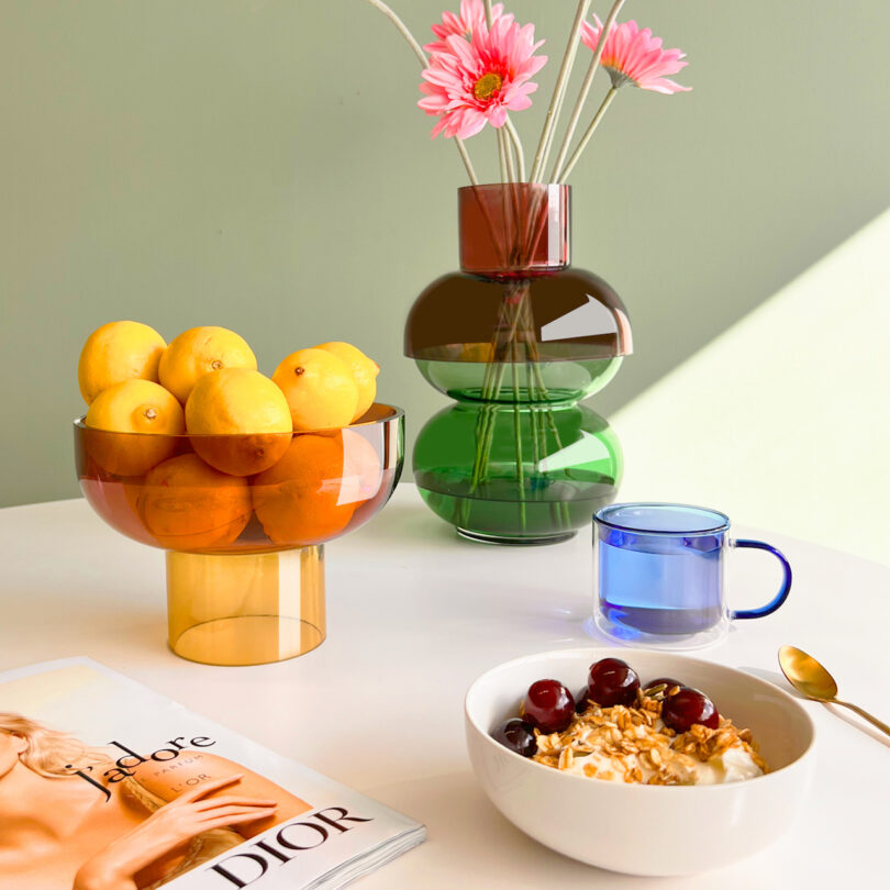 A bowl of cereal with cherries and nuts, a blue mug, a vase with pink flowers, a bowl of lemons and oranges, and a magazine are on a white table