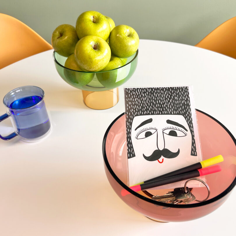 A table with a bowl of green apples, a clear blue mug, and a pink bowl containing a drawing of a face with a mustache, along with two markers and a set of keys