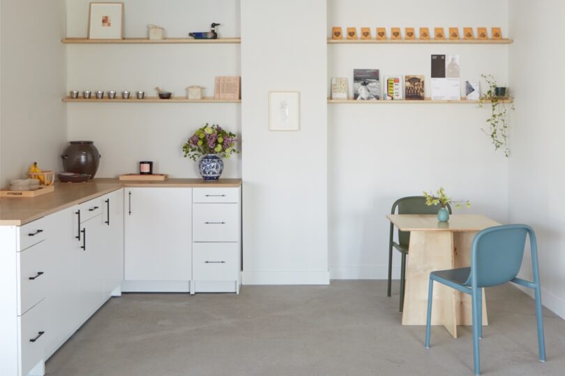 A minimalist kitchen and dining area with white cabinets, wooden countertop, shelves with decor and books, a small table with two chairs, and a vase with flowers