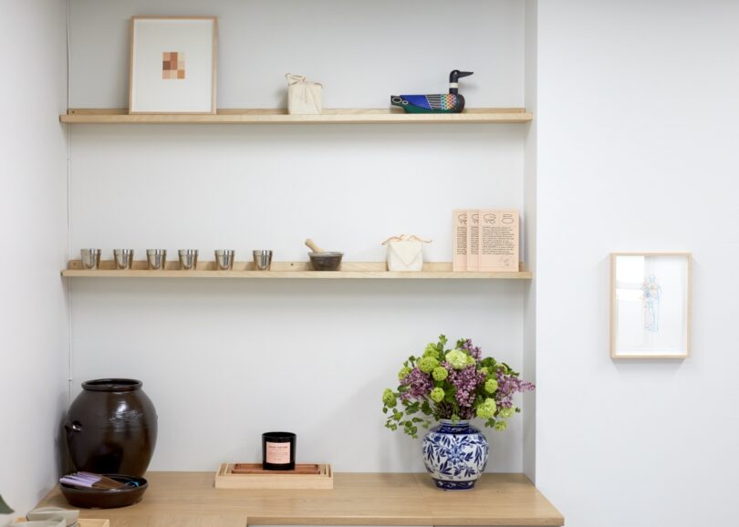 A minimalist shelf setup includes small metal cups, a mortar, and a wooden figure on the left. A vase with flowers and a candle sit on the lower wooden counter. Artwork and small items are on adjacent shelves