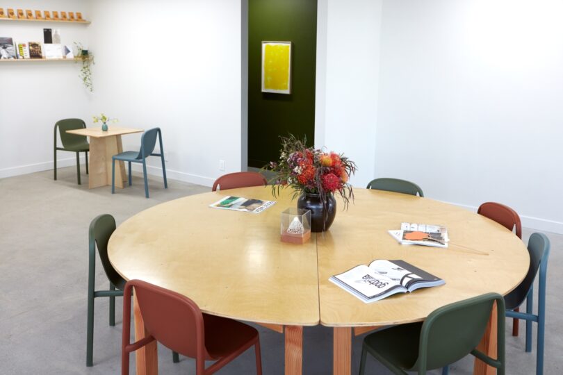 A modern room with a round wooden table, surrounded by chairs, featuring books, magazines, and a vase of flowers. In the background, there's a small table with chairs, bookshelves, and wall art