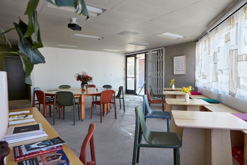 A room with tables and chairs, books and flowers on a shelf, a large window with sheer curtains, and a whiteboard