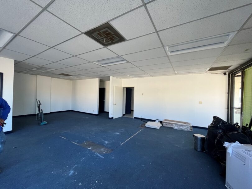 A partially renovated office room with white walls, blue carpet, and drop ceiling tiles. Some cleaning equipment and bags are on the floor. The room has open and closed doors leading to other areas