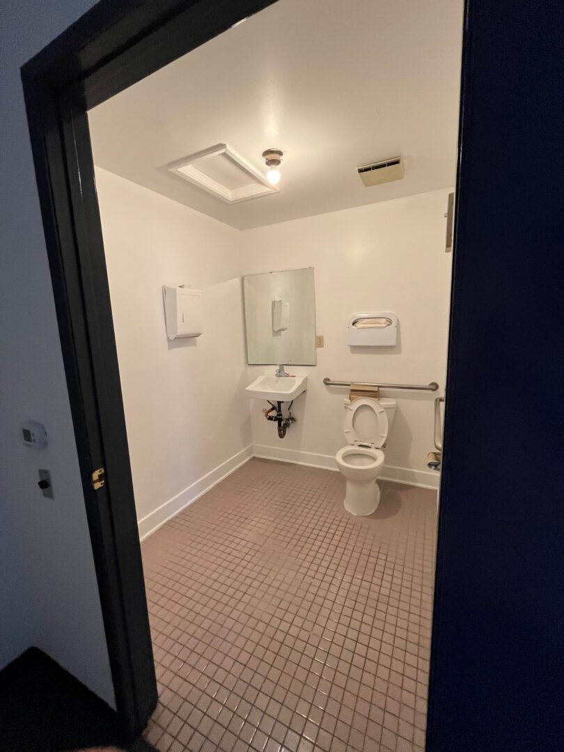 A small, clean restroom with white walls, a tiled floor, a sink under a mirror, a toilet with a safety rail, and a paper towel dispenser