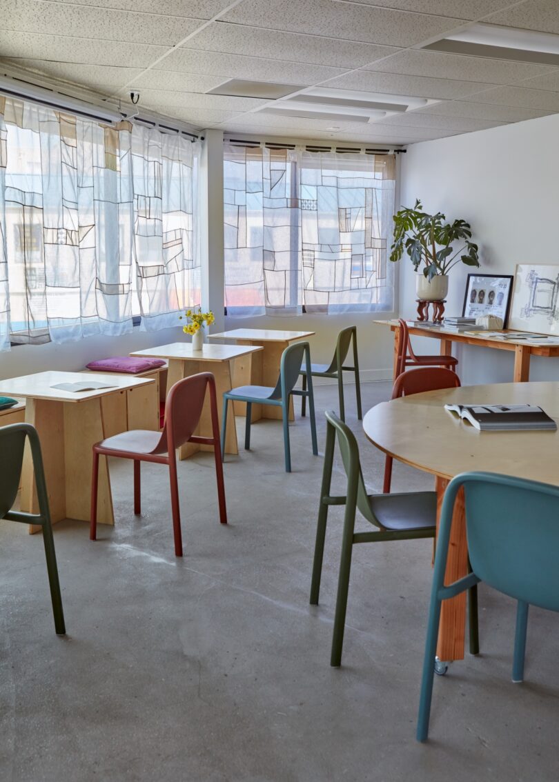 A bright, modern classroom with various tables and colorful chairs. Large windows with sheer curtains let in natural light. A plant, books, and a framed picture decorate the space