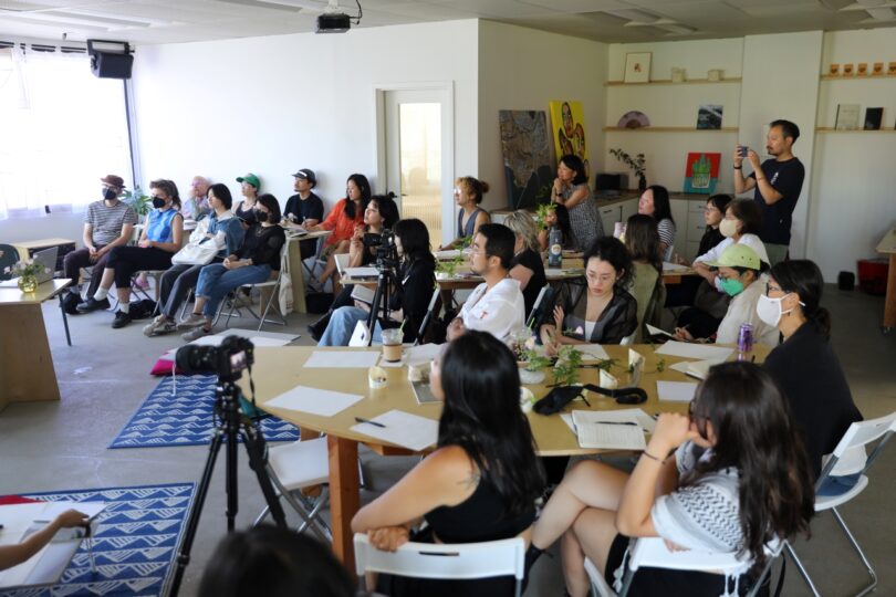 A group of people seated in a room attentively listen to a presentation. Some are taking notes while one person stands at the back holding a camera. A tripod is set up at the front