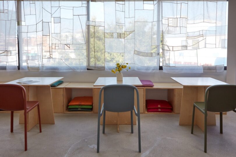 A minimalist office with three tables, each with a different colored chair and a vase of yellow flowers on the middle desk. The window has translucent geometric-patterned curtains