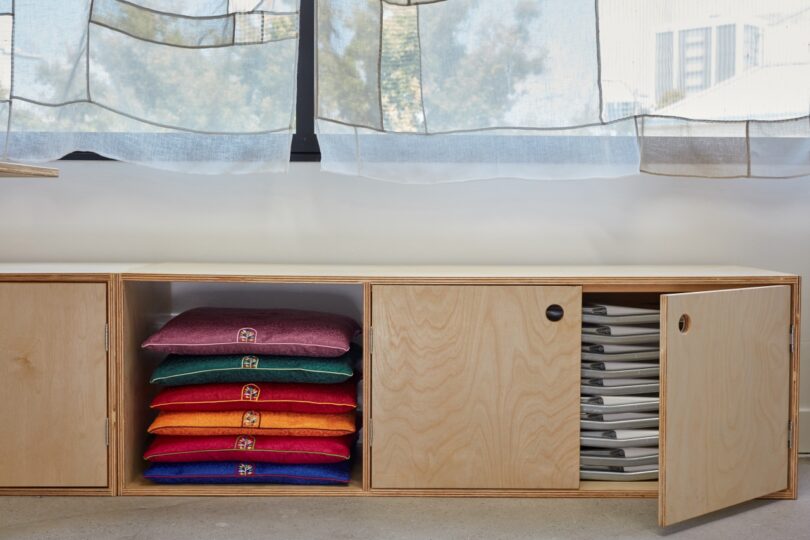 A wooden storage unit with open doors revealing neatly stacked colorful cushions on the left and white folded items on the right. Light filters through sheer curtains in the background