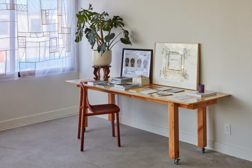 A wooden desk holds books, papers, framed artwork, and a potted plant. A red chair is positioned in front of the desk. A window with semi-transparent curtains is on the left.