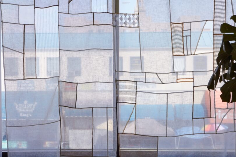 Windows covered with sheer curtains featuring a geometric pattern of squares and rectangles. Sunlight filters through, illuminating the room softly. A partial view of buildings outside is visible