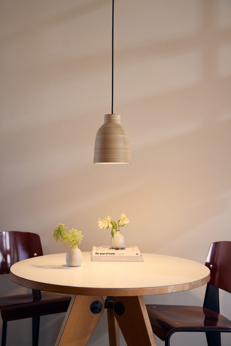 A minimalist round table with two small flower vases on top and two chairs on either side, illuminated by a pendant light hanging from the ceiling
