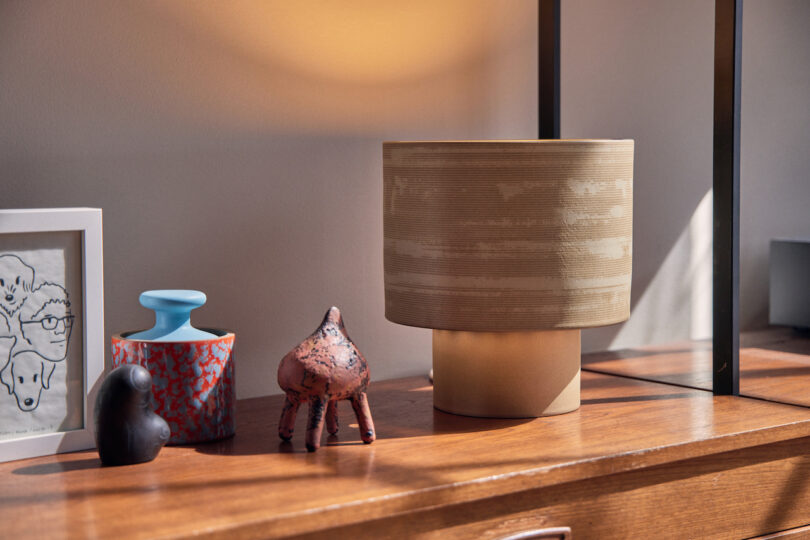 A wooden table with a framed drawing, a blue and red jar, a small abstract horse sculpture, a dark elephant figurine, and a beige lamp
