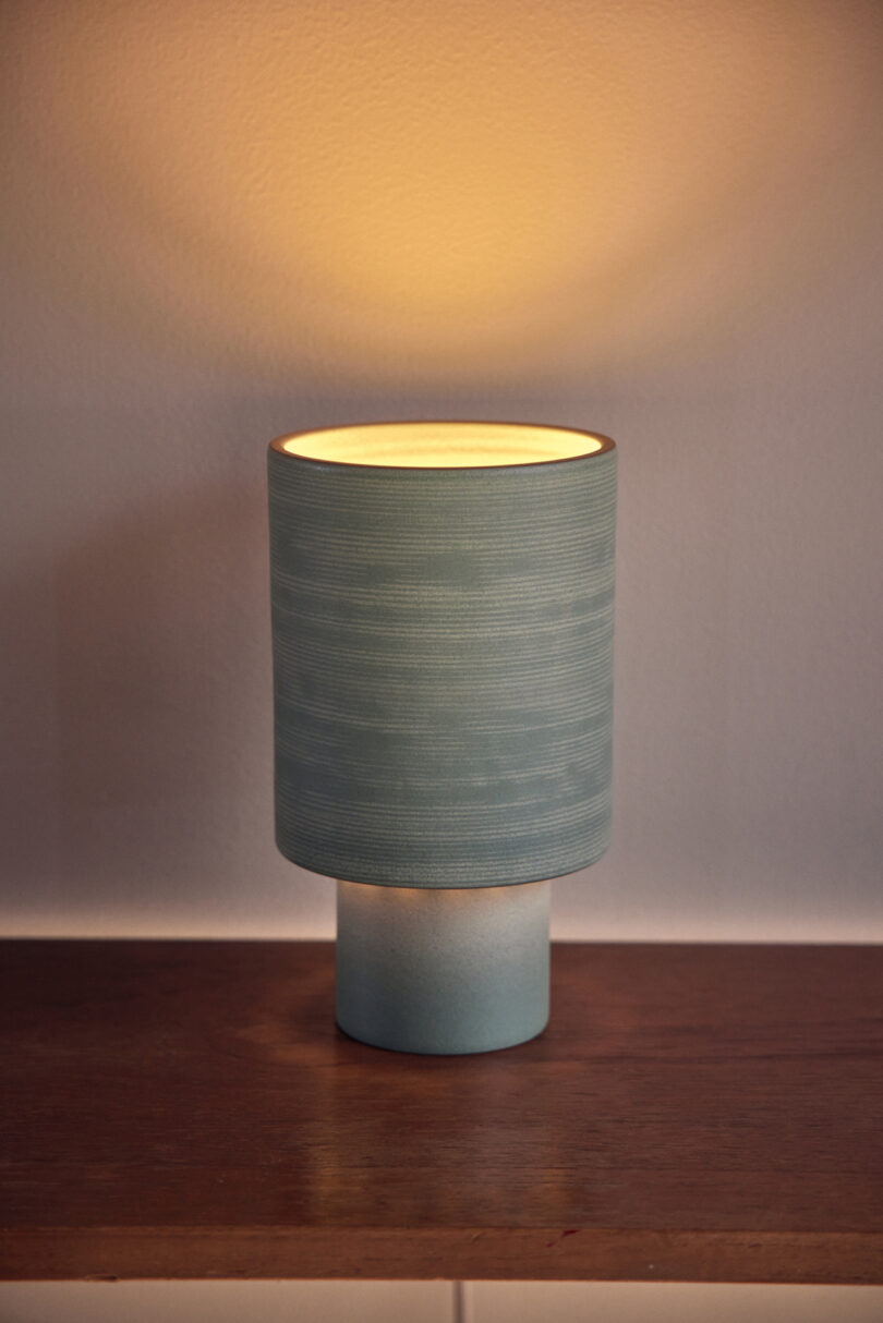 A small, cylindrical table lamp with a blue shade emits a soft, warm light while placed on a wooden surface against a plain wall