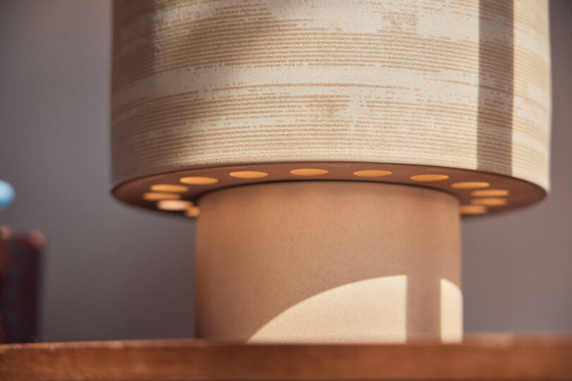 Close-up view of a beige cylindrical table lamp with a textured lampshade, lit from within, casting light through perforated holes at the base