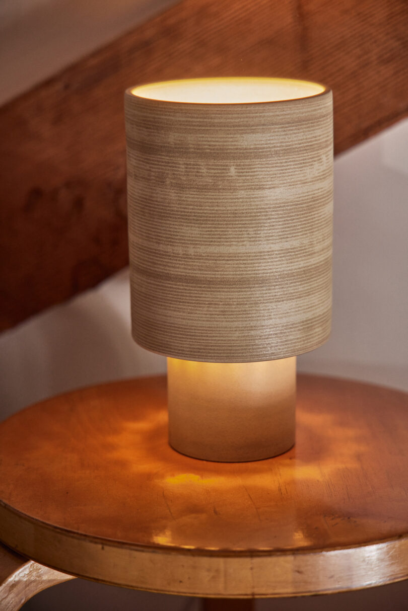 A cylindrical table lamp with a beige, textured shade sits on a wooden surface, illuminated and casting a warm glow