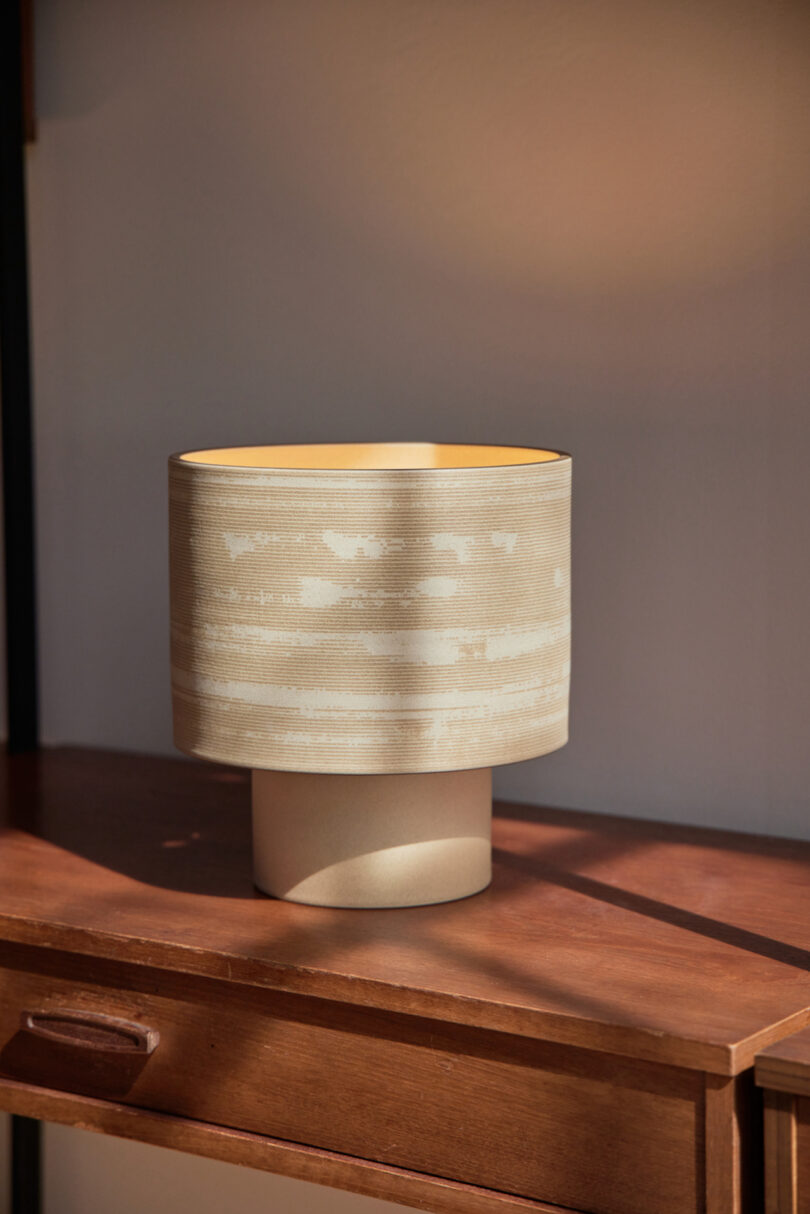 A beige ceramic lamp with a textured cylindrical shade sits on a wooden table, illuminated by natural sunlight