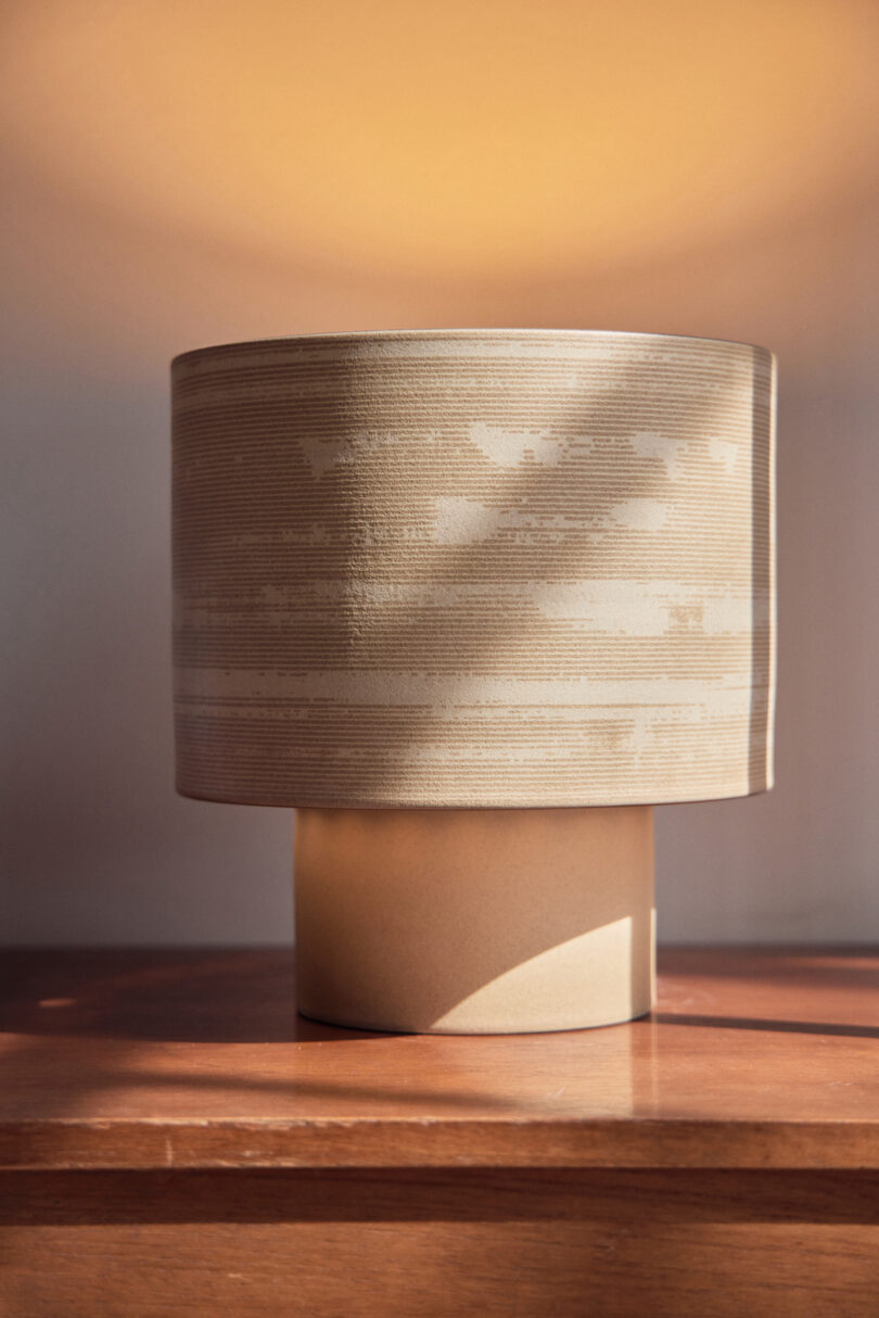 A beige, textured cylindrical lamp sits on a wooden surface with soft light casting shadows behind it