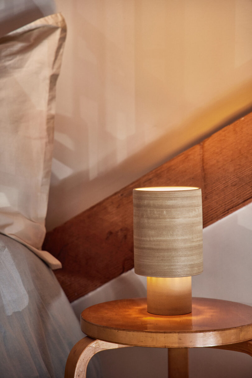 A small, cylindrical bedside lamp with a wooden finish is placed on a round wooden table next to a bed with a white pillow