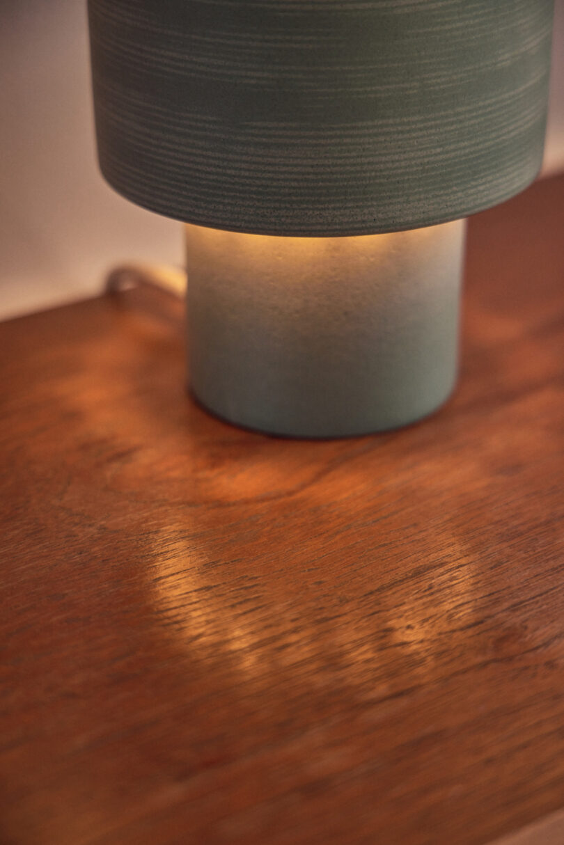 Close-up of a lit table lamp with a cylindrical base and shade, casting a warm light on a wooden surface