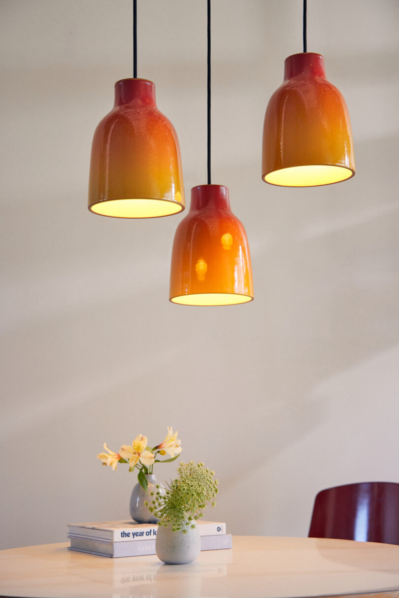 Three orange pendant lights hang above a round, white table with a small vase of flowers and two stacked books