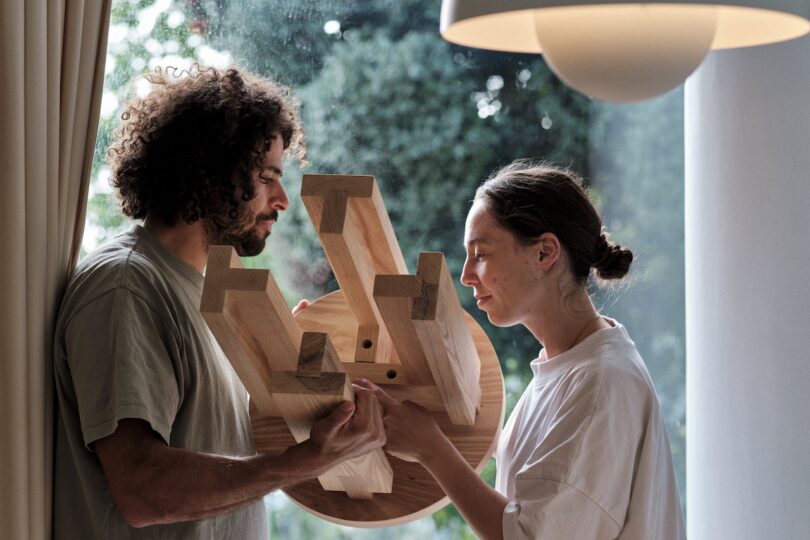 Two people are holding and examining a wooden piece of furniture near a large window with greenery outside. A round, white light fixture is visible above