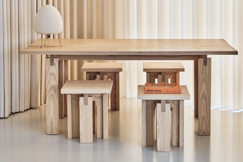 A wooden table with four matching wooden stools. A small, round lamp is on the table alongside an open orange book. Beige curtains in the background