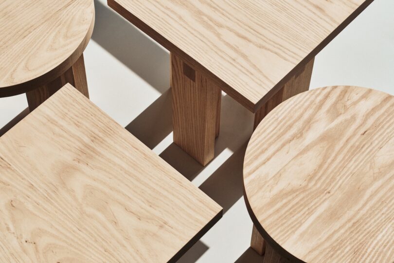 Three wooden tables arranged together, two with round tops and one with a square top, are displayed on a light surface