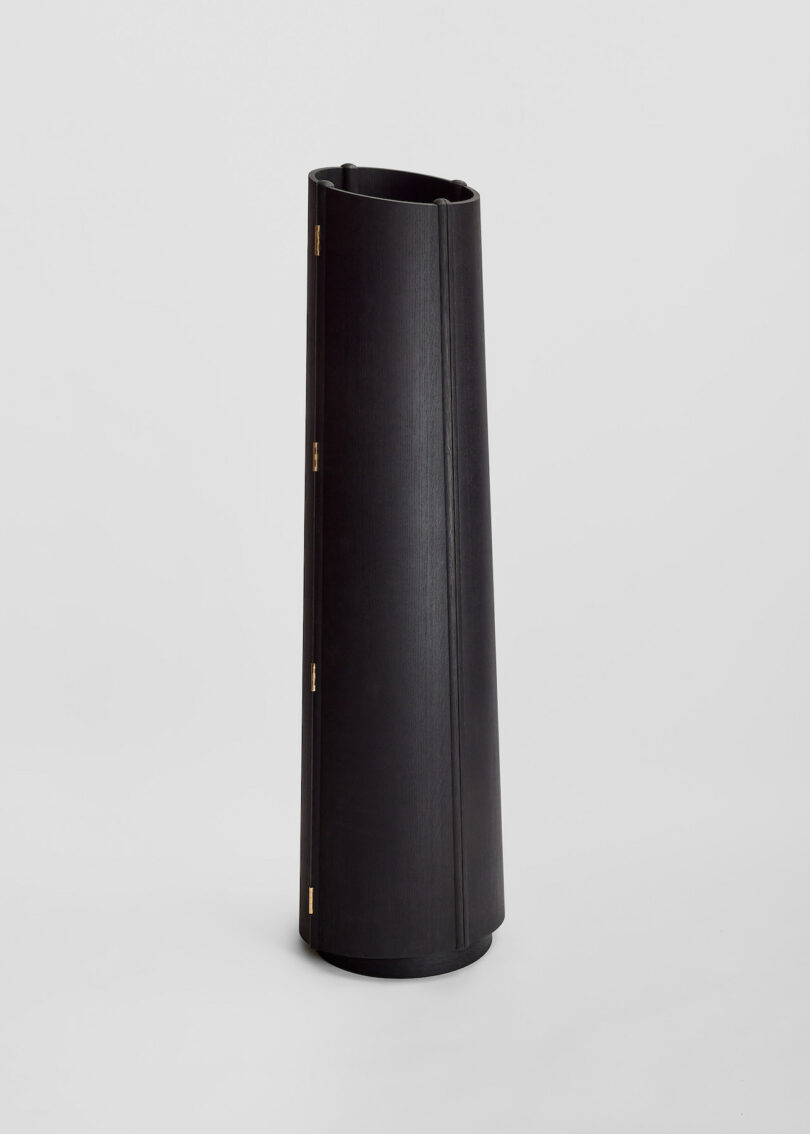 A tall, black vase with a cylindrical shape and minimalist design stands upright on a plain background.