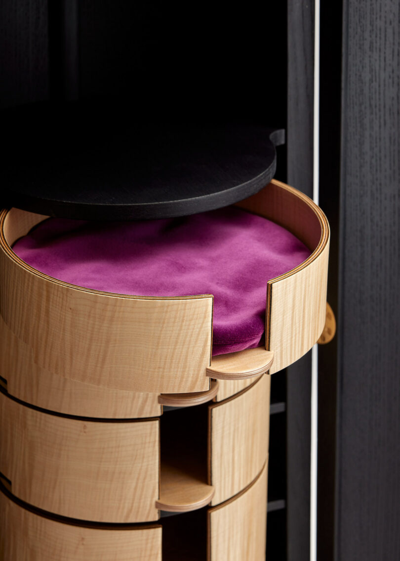 A wooden cylindrical cabinet with a circular black lid and purple velvet cushion on top, featuring multiple drawers.