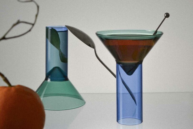 Two modern, colorful glasses are on a surface. One glass has a tall, conical design, while the other is inverted, resembling a martini glass. There is an orange object and a twig in the foreground.