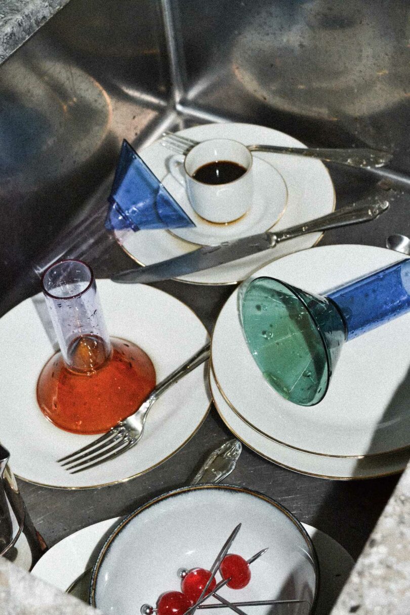 A kitchen sink filled with unwashed dishes, including plates, cutlery, colored glass cups, a coffee cup, and a small plate with skewered cherries