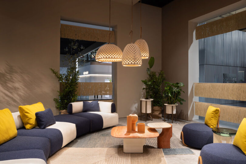 Modern living room with a circular arrangement of blue and white sofas, yellow cushions, three hanging pendant lights, and a central coffee table. Neutral tones and plants enhance the cozy atmosphere