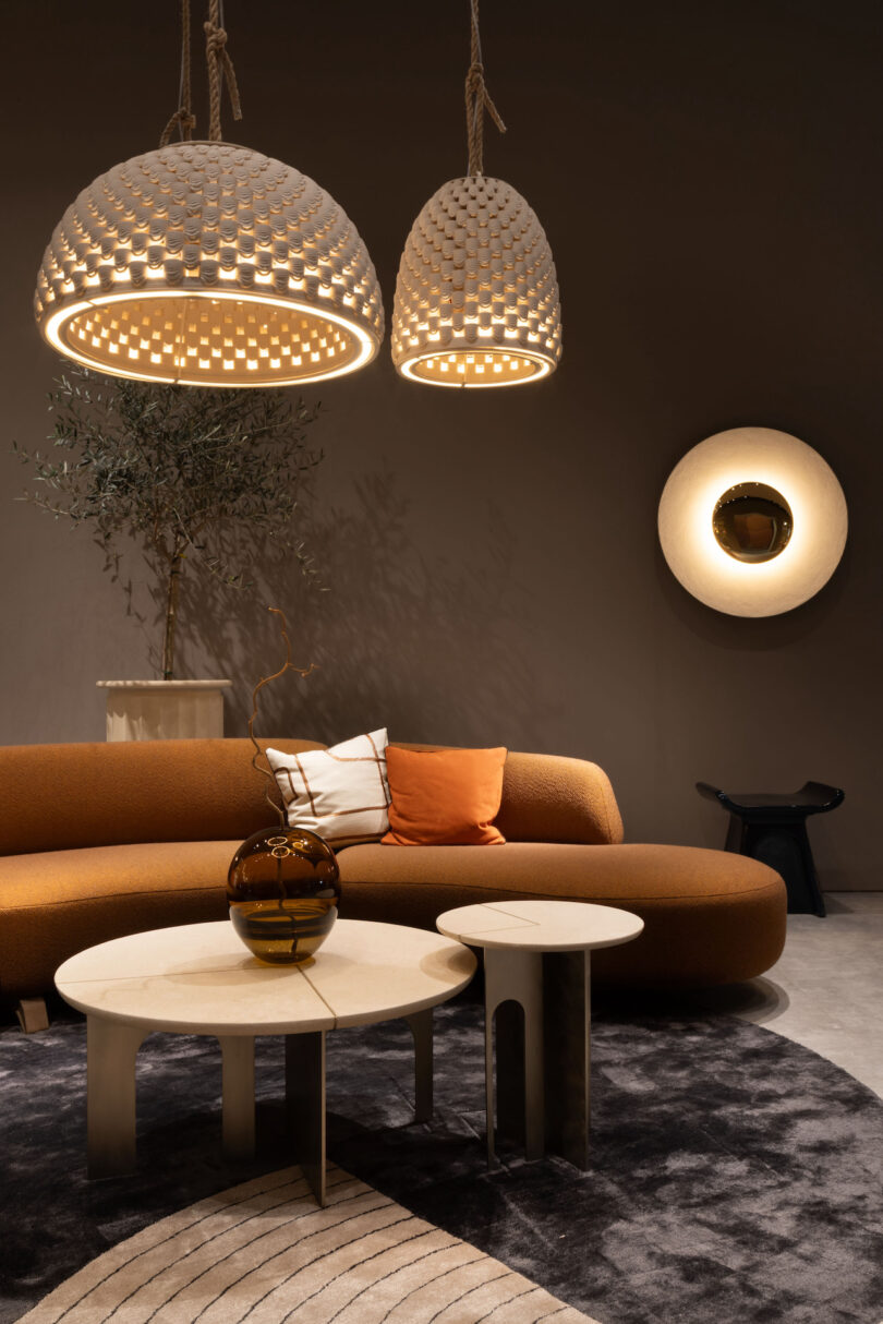 Modern living room featuring a brown sectional sofa, a round coffee table with a decorative vase, two hanging light fixtures, a wall-mounted lamp, and a plant in the background