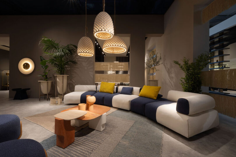 A modern living room features a large, curved sectional sofa in white and navy blue, complemented by wooden coffee tables, hanging pendant lights, and various plants